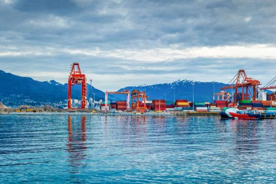 A panoramic view of a major Canadian shipping port with industrial cranes, colorful shipping containers, and cargo vessels set against a backdrop of snow-capped mountains and a dramatic sky.